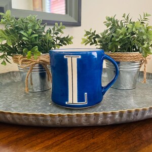 May include: A cobalt blue ceramic mug with a white letter "L" is centered in the image. The mug is on a galvanized metal tray, with two small potted plants on either side. The plants are in small metal buckets wrapped with twine.