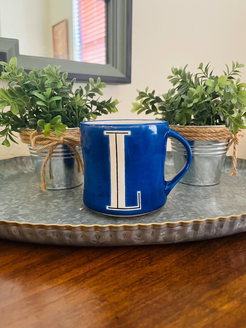 May include: A cobalt blue ceramic mug with a white letter "L" is centered in the image. The mug is on a galvanized metal tray, with two small potted plants on either side. The plants are in small metal buckets wrapped with twine.
