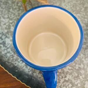 May include: A ceramic mug with a blue rim and handle, and a cream-colored interior. The mug is empty and sits on a textured surface. The mug's handle is a solid blue color.