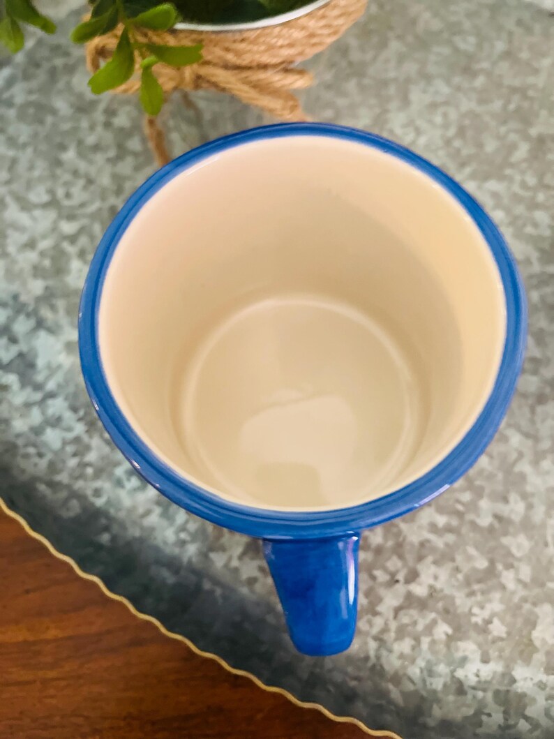 May include: A ceramic mug with a blue rim and handle, and a cream-colored interior. The mug is empty and sits on a textured surface. The mug's handle is a solid blue color.