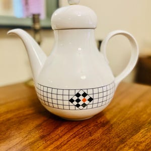 May include: White ceramic teapot with a lid and handle, featuring a black and white checkered pattern with a red and black diamond design. The teapot is set on a wooden surface.