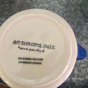 May include: A white ceramic mug with a blue handle and rim. The bottom of the mug features the text "ANTHROPOLOGIE hand painted". The mug is dishwasher and microwave safe, made in China.
