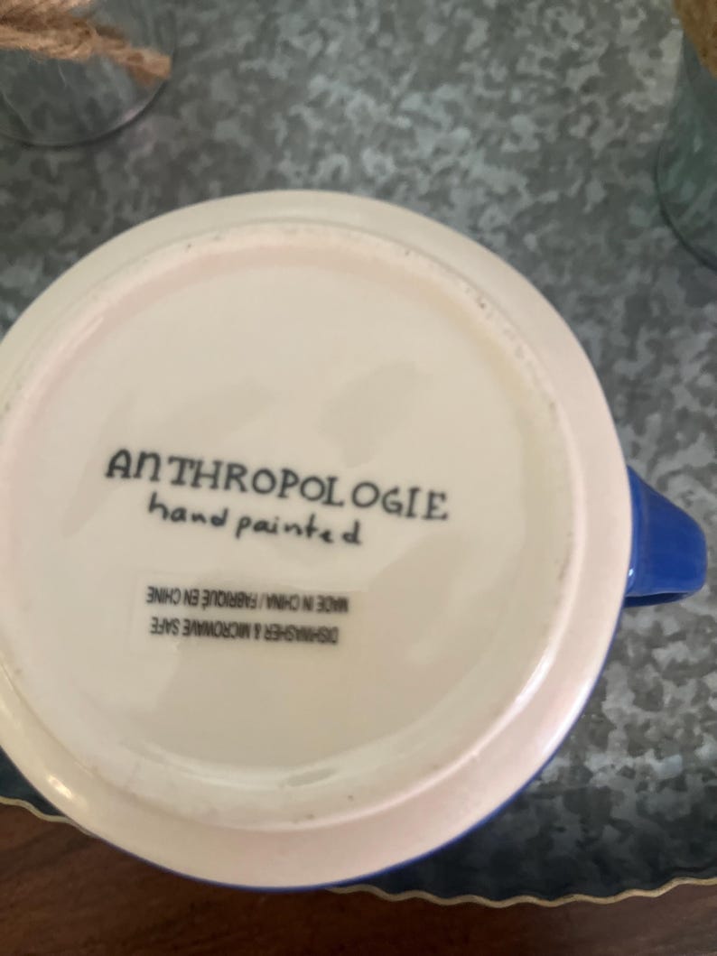 May include: A white ceramic mug with a blue handle and rim. The bottom of the mug features the text "ANTHROPOLOGIE hand painted". The mug is dishwasher and microwave safe, made in China.