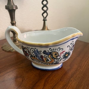 May include: White ceramic gravy boat with a blue and gold rim and a hand-painted design of a yellow bird with red, blue, and green accents.