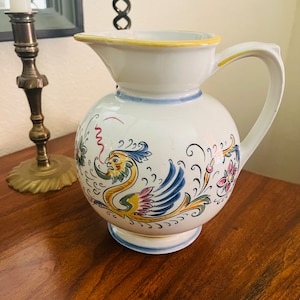 May include: A white ceramic pitcher with a round body and a handle. The pitcher is decorated with a colorful bird and floral design. The rim is yellow, and the base is blue. A vintage decorative piece.