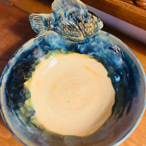 May include: A ceramic bowl with a blue, green, and yellow glaze. The bowl has a fish-shaped handle.