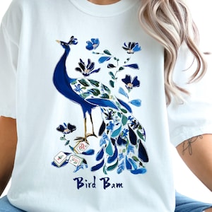 May include: White t-shirt featuring a blue peacock design with floral accents. The peacock has a detailed tail and is surrounded by blue flowers. The text "Bird Bam" is printed in a script font below the peacock.
