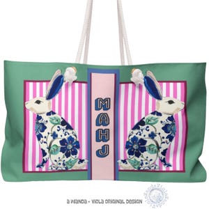 May include: A large tote bag with a green background and white rope handles. The bag features two rabbit illustrations with blue floral patterns. The center has pink and white stripes with the word "MAHJ" in blue. The design is framed in a dark pink border.