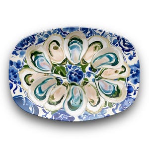 May include: An oval serving platter featuring a painted pattern of multiple oyster shells in shades of cream, teal, and dark blue, surrounded by a decorative border of blue and white floral motifs on a white background.
