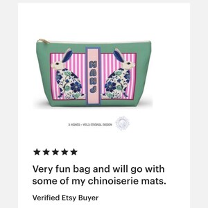 May include: A teal cosmetic bag with a gold zipper and a rectangular design featuring two rabbits with blue and white floral patterns. The bag has a pink and white striped center panel with the letters "MAHJ".