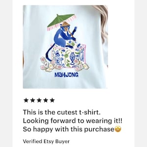 K&ouml;nnte beinhalten: Wei&szlig;es T-Shirt mit einem blauen Affen-Design, der einen gr&uuml;nen Sonnenschirm h&auml;lt, umgeben von Mahjong-Steinen. Das Wort "MAHJONG" ist blau gedruckt. Das T-Shirt ist hellblau.