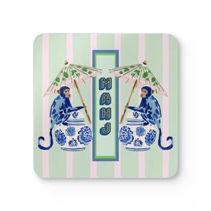 Op de afbeelding: Een vierkante onderzetter met een lichtgroene en roze gestreepte achtergrond. Twee blauwe apen zitten op blauw-witte gemberpotten en houden elk een roze en groene parasol vast. Een centrale groene rechthoek bevat de blauwe tekst MAHJ.
