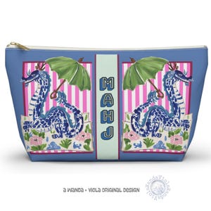 May include: Blue cosmetic bag with a gold zip. The bag features a design with blue and white dragons, green umbrellas, and the word "MAHJ" in blue letters. Pink and white striped background with floral accents.