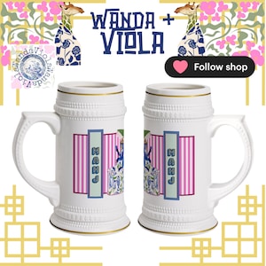 May include: Two white ceramic beer steins with gold trim and handles. Each stein features a design with pink stripes, blue and green accents, and the word "MAHJ". The top of the image has the text "WANDA + VIOLA" and giraffe illustrations.