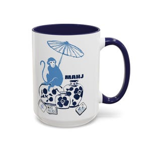 May include: A white ceramic mug with a dark blue interior and handle. The exterior features a blue graphic illustration of a monkey holding an umbrella, sitting on a patterned vase, with additional vases and mahjong tiles and the text MAHJ.