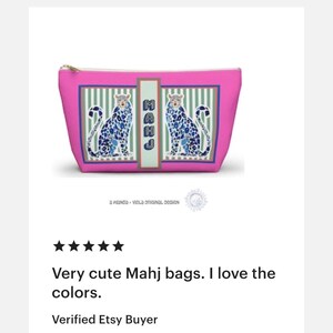 Puede incluir: Un neceser rosa con cremallera dorada, con un dise&ntilde;o de dos criaturas azules parecidas a leopardos. La bolsa tiene un panel rectangular con la palabra "MAHJ" verticalmente. El fondo tiene rayas verdes y blancas.