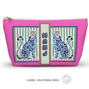 Op de afbeelding: Een roze make-uptas met twee gestileerde blauwe en witte luipaardontwerpen. De tas heeft een groen-witte gestreepte achtergrond, een blauw-groene rand en het woord "MAHJ" in blauw. De tekst "a Wanda + Viola Original Design" staat onderaan.