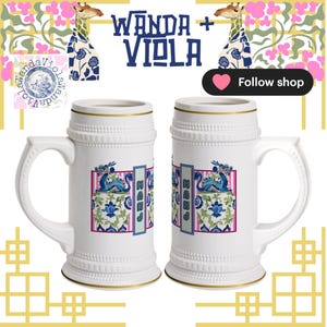 May include: Two white ceramic beer steins with gold trim. Each stein features a decorative panel with blue and white floral designs and a blue dragon. The text "WANDA + VIOLA" is above the steins.