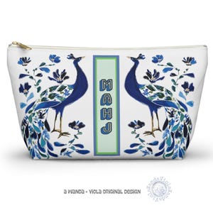 May include: A white cosmetic bag with a gold zip, decorated with two blue peacocks and floral patterns. The bag has the word "MAHJ" in a vertical green rectangle. The text "A Wanda + Viola Original Design" is printed at the bottom.