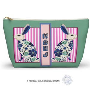 May include: A green zippered pouch featuring two rabbit illustrations with blue floral patterns. The pouch has a pink and white striped background and the word "MAHJ" in blue. The design is by Wanda + Viola.