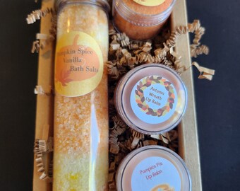 May include: A gift set featuring fall-themed bath and body products. The set includes a glass tube of pumpkin spice vanilla bath salts, a lip scrub, and two lip balms. The products are in a brown box with decorative paper.