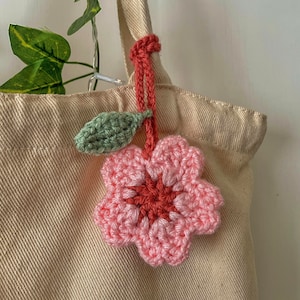 May include: A crocheted pink flower with a green leaf attached to a red string. The flower is hanging from a beige tote bag.