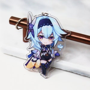 May include: A clear acrylic keychain featuring a cartoon anime character with blue hair, a white dress, and a blue and white flower on her head. The character is holding a blue and white flower in her right hand.