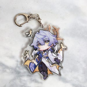 May include: A clear acrylic keychain featuring a chibi-style anime character with silver hair, yellow eyes, and a white and blue outfit. The keychain has a silver clasp and several small, stylized leaf-like accents.