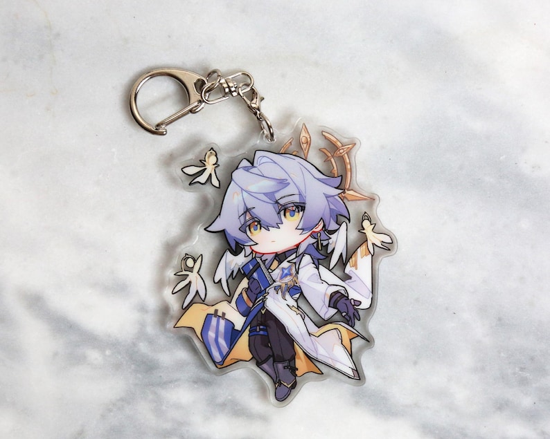 May include: A clear acrylic keychain featuring a chibi-style anime character with silver hair, yellow eyes, and a white and blue outfit. The keychain has a silver clasp and several small, stylized leaf-like accents.