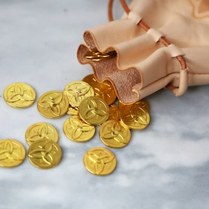 May include: A small, light brown leather pouch with a drawstring closure is spilling out gold-colored coins with a Celtic knot design.