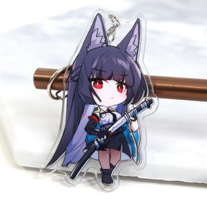 Op de afbeelding: Een transparante acryl sleutelhanger met een anime-personage met zwart haar, rode ogen en een blauw-witte outfit. Het personage houdt een zwaard vast en heeft wolvenoren.
