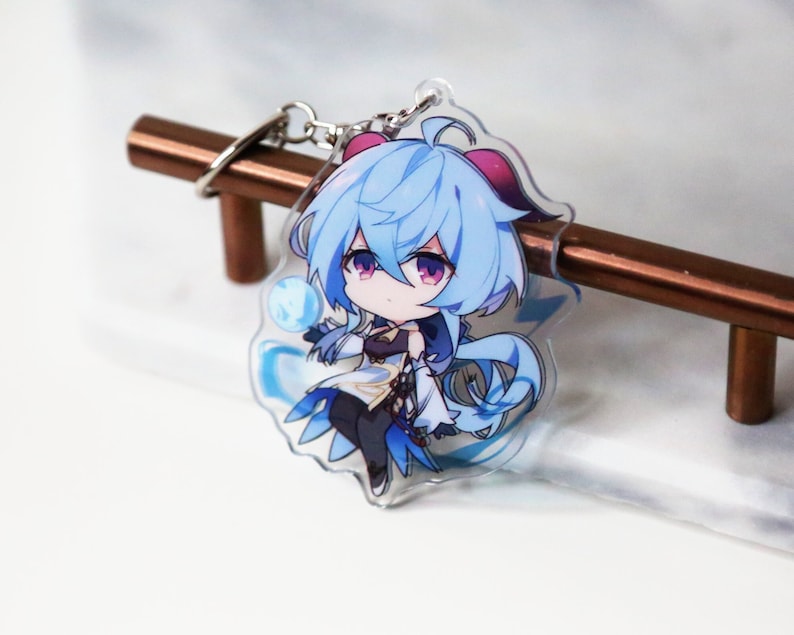 May include: A blue and white anime character keychain with a blue and white background. The character is a young woman with long blue hair and a white dress. She is holding a blue orb in her hand.