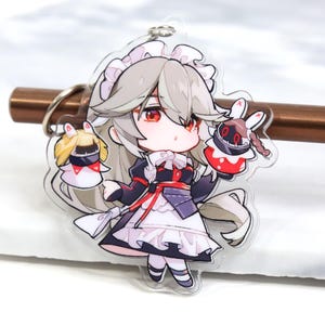 Op de afbeelding: Een anime-personage met wit haar en rode ogen, gekleed in een zwart-witte dienstmeidenoutfit en een rood-zwart knuffelbeest vasthoudend. Het personage houdt een tweede knuffelbeest vast met een wit en rood ontwerp. Het personage is een sleutelhanger.