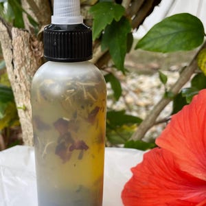 May include: A clear glass bottle with a black cap and a dropper top. The bottle is filled with a yellow liquid and contains dried flowers and herbs. The bottle is sitting on a white surface next to a red hibiscus flower.
