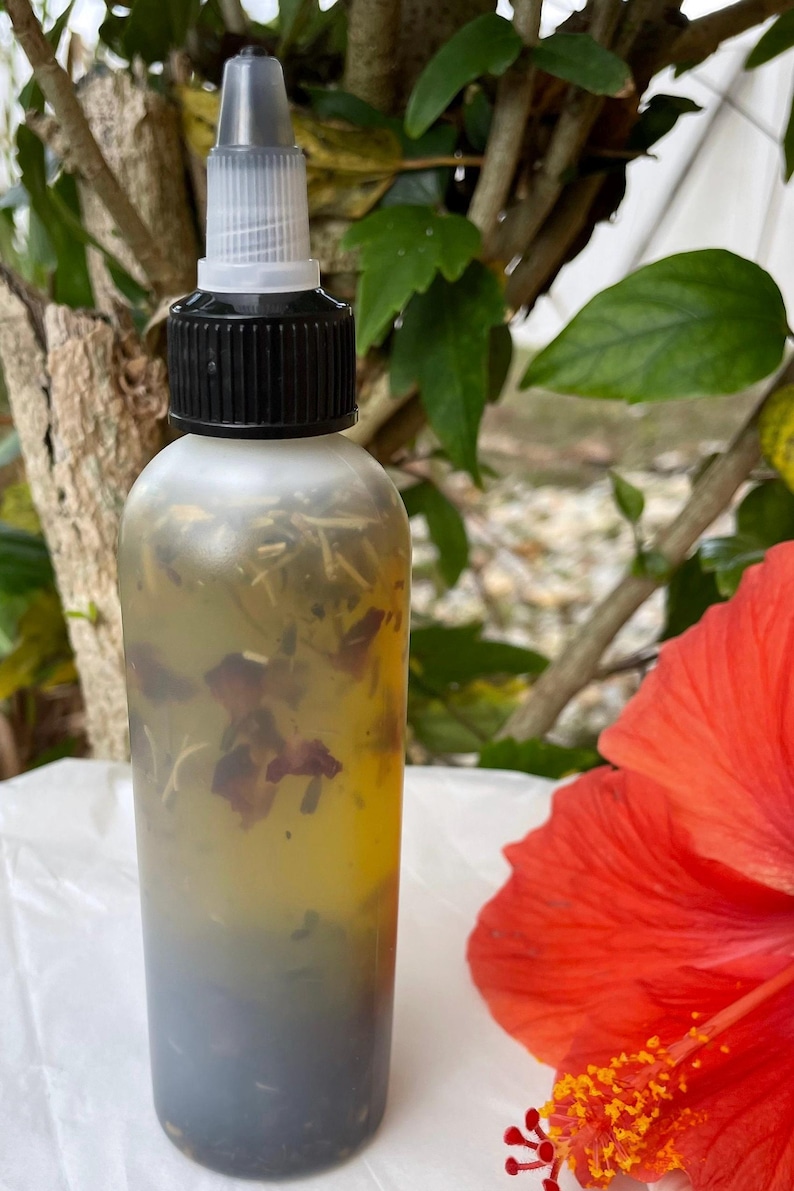 May include: A clear glass bottle with a black cap and a dropper top. The bottle is filled with a yellow liquid and contains dried flowers and herbs. The bottle is sitting on a white surface next to a red hibiscus flower.