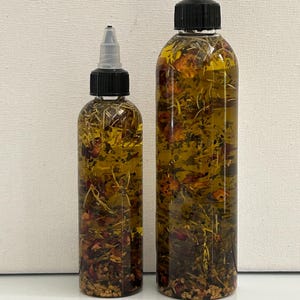 May include: Two clear glass bottles with black caps and droppers filled with a golden oil infused with dried herbs and flowers.