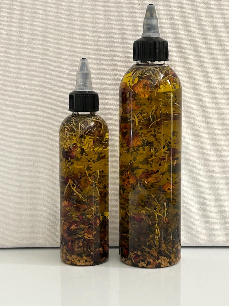 May include: Two clear glass bottles with black caps and droppers filled with a golden oil infused with dried herbs and flowers.