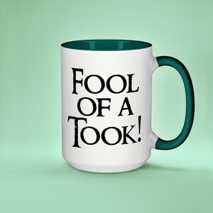 May include: White ceramic mug with a green rim and handle. The mug has black text that reads "Fool of a Took!"
