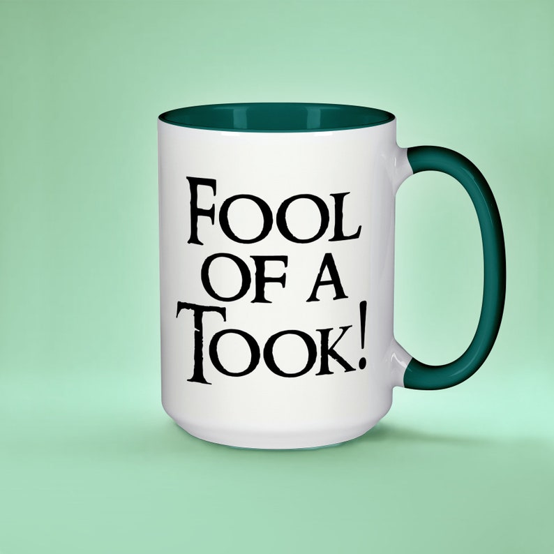 May include: White ceramic mug with a green rim and handle. The mug has black text that reads "Fool of a Took!"