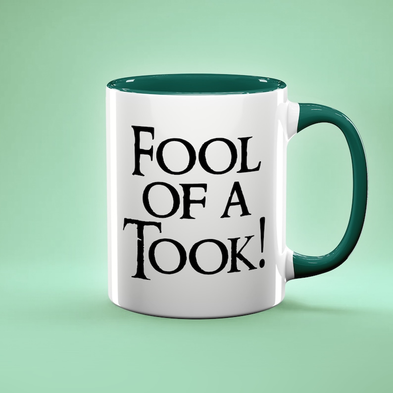 May include: White ceramic mug with a green rim and handle. The mug has black text that reads "Fool of a Took!"