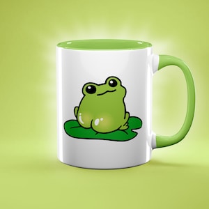 May include: White ceramic mug with a green interior and handle. The mug features a cartoon frog sitting on a lily pad. The frog is green with large black eyes and a smiling expression. The background is a light green.