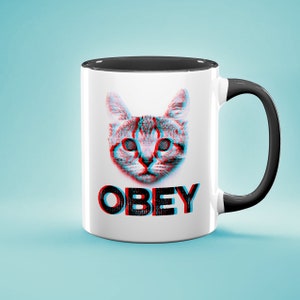May include: White ceramic mug with a black rim and handle. The mug features a 3D image of a cat with swirling eyes and the word "OBEY" printed below the cat.