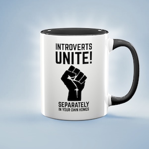 Puede incluir: Taza de cerámica blanca con interior y asa negros. La taza presenta el texto "INTROVERTS UNITE!" encima de un gráfico de un puño negro, con la frase "SEPARATELY IN YOUR OWN HOMES" debajo. La taza está sobre un fondo azul claro.