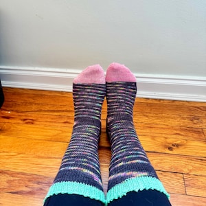 May include: Hand-knitted socks with pink toes, a dark grey body with rainbow stripes, and a light green scalloped cuff. The socks are worn over dark blue leggings. The background shows a wooden floor and a white wall.