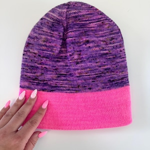 May include: A knitted beanie hat with a vibrant color scheme. The top portion features a speckled pattern in shades of purple, pink, and brown, while the bottom band is a solid bright pink. The hat is made of soft yarn.
