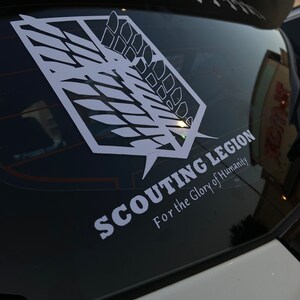 Op de afbeelding: Een witte vinyl sticker op een zwart autovenster. De sticker heeft een gestileerd vleugelontwerp met de tekst "SCOUTING LEGION" en "For the Glory of Humanity".
