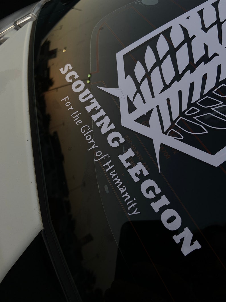 Op de afbeelding: Een witte sticker op een autoruit met de tekst "SCOUTING LEGION For the Glory of Humanity" en een gestileerd vleugelontwerp.