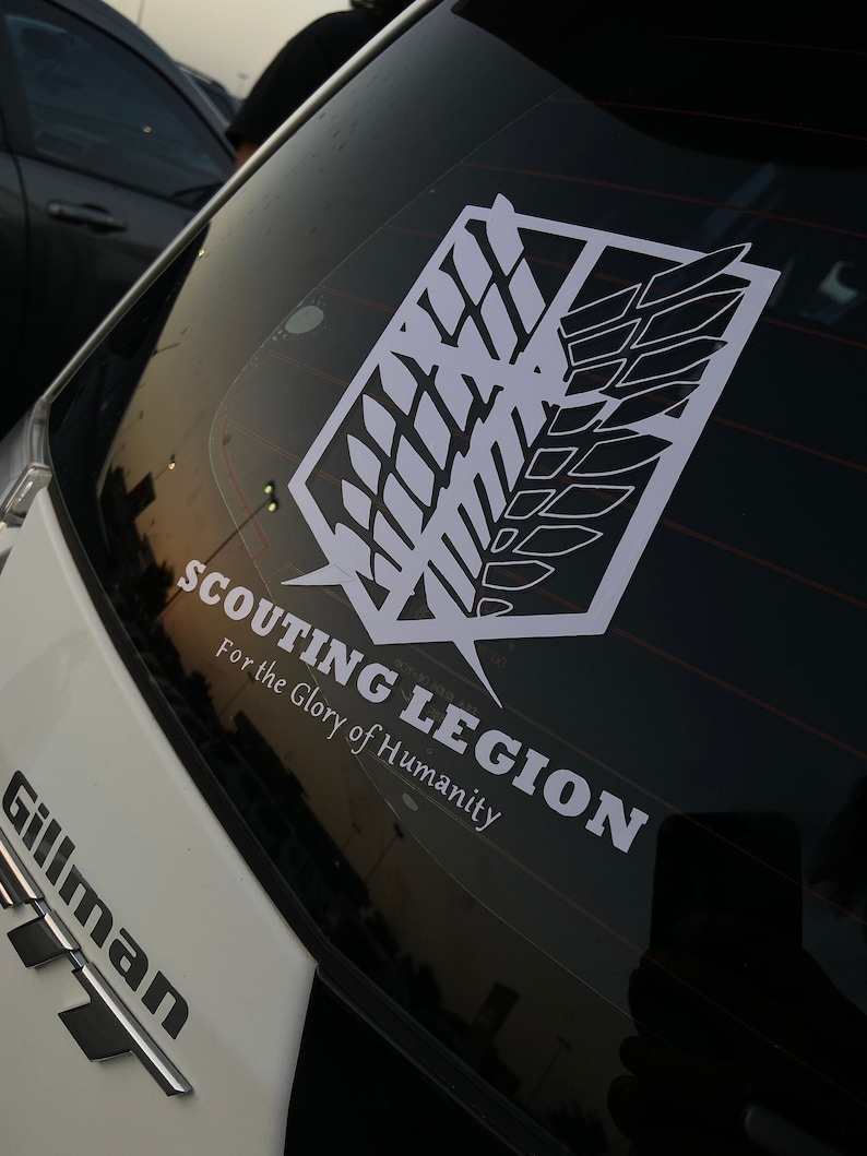 Op de afbeelding: Een witte auto met een zwart-witte sticker op de achterruit. De sticker is een gestileerde afbeelding van een vleugel met de tekst "SCOUTING LEGION For the Glory of Humanity".