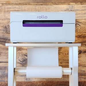 May include: White label printer with a purple stripe and the word "rollo" on the front, sitting on a white metal stand with a roll of white paper.