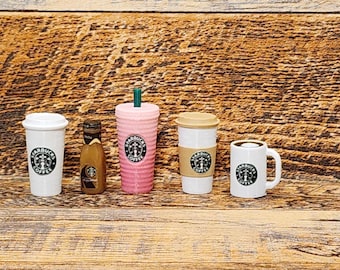 May include: Five Starbucks cups and a bottle of creamer. The cups are white with a black Starbucks logo and a brown lid. The creamer bottle is brown with a white Starbucks logo. The cups are arranged in a row on a wooden surface.
